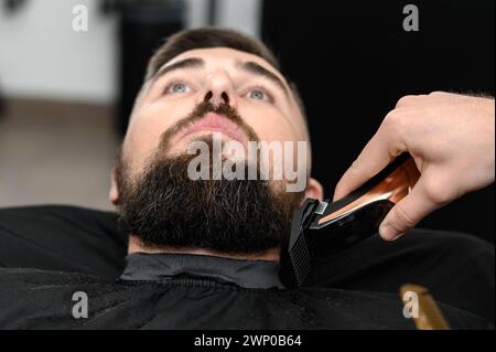 Tagliare la barba a un gentiluomo in un barbiere con un regolacapelli. Accorciare la lunghezza della barba dai lati dal maestro per il cliente. Foto Stock