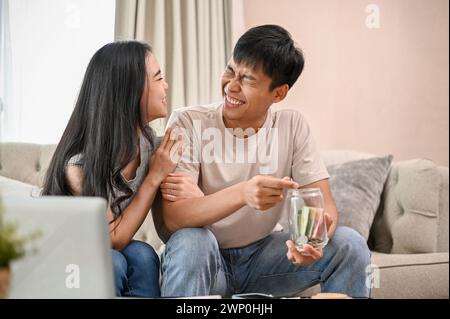 Una giovane coppia asiatica, felice e felice, sta progettando i loro risparmi insieme in salotto, mettendo monete e un po' di soldi in un barattolo. Foto Stock