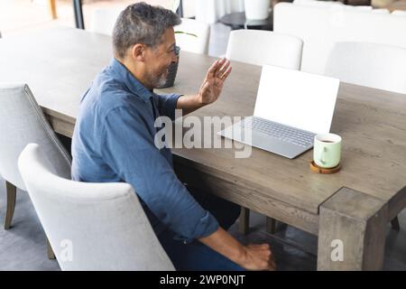 Un anziano birazziale con una camicia blu ondeggia durante una videochiamata sul suo portatile Foto Stock