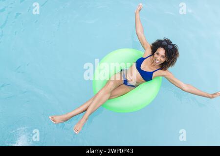 Donna birazziale ama galleggiare in piscina su un tubo interno verde, sorridendo alla telecamera Foto Stock
