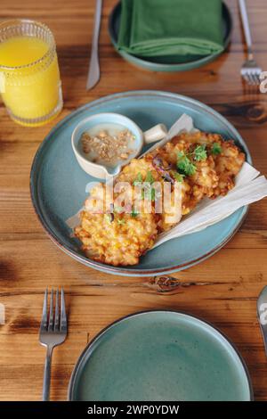 Un piatto di frittura con un lato di salsa da immersione si trova su un tavolo di legno. Il tavolo è allestito con una forchetta, un coltello, un cucchiaio e un bicchiere di succo d'arancia Foto Stock