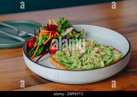 Un piatto di toast all'avocado con un lato di insalata. Il piatto è su un tavolo di legno. L'insalata è composta da lattuga, carote e pomodori Foto Stock