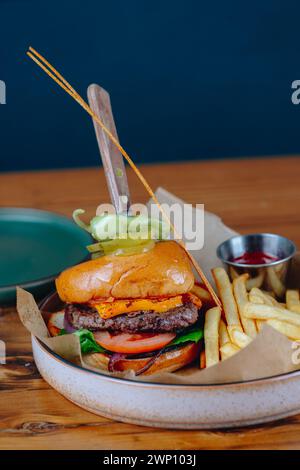 Un hamburger con una lancia di sottaceti sopra e un lato di patatine fritte. L'hamburger viene servito su un piatto bianco e le patatine fritte sono in un piccolo recipiente. La scena è casua Foto Stock