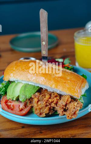 Un panino con un coltello che fuoriesce è su un piatto blu. Il panino è ricoperto di lattuga, pomodori e avocado. C'è anche un bicchiere di o. Foto Stock