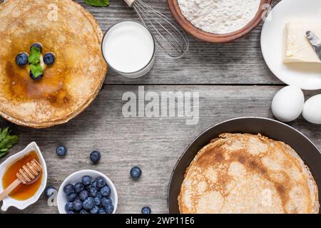 Pila di pancake sottili con mirtilli e miele e padella con deliziose crepe e ingredienti per cucinare su un rustico tavolo in legno. Vista dall'alto Foto Stock