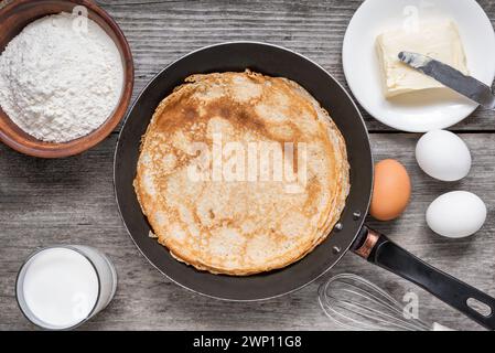 Padella con deliziose crepe e ingredienti per cucinare su un rustico tavolo di legno. Pancake sottili in una padella con farina, uova, latte e burro. Superiore Foto Stock