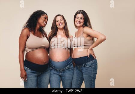 Si aspettano madri con background diversi che si trovino insieme in uno studio, mostrando i loro piccoli dossi. Indossano jeans e reggiseni, sorridendo mentre sostengono Foto Stock