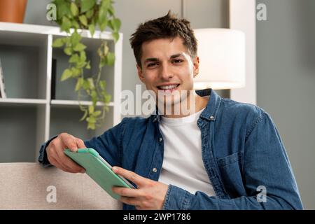 Uomo sorridente e rilassato con un tablet digitale in un appartamento moderno. Bel giovane spensierato che lavora con il touchpad seduto sul divano a casa Foto Stock