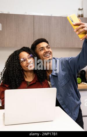 Giovane uomo e donna adulta che fanno una foto nel loro appartamento usando un computer. Coppia sorridente felice e diversificata che scatta un selfie mentre lavora online con Foto Stock