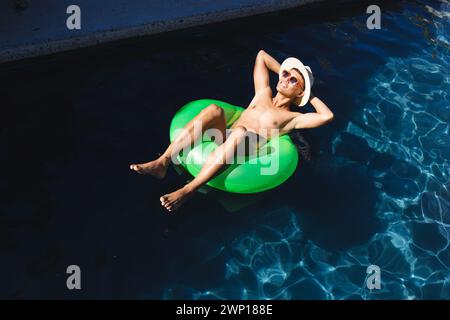 Un giovane asiatico si rilassa su un galleggiante verde in una piscina, indossando un cappello bianco e occhiali da sole Foto Stock