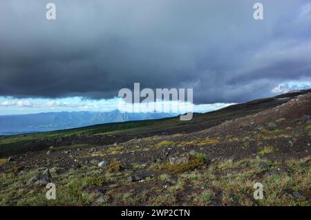 Pendenza scura del vulcano Villarrica con vegetazione sparsa, nuvole scure e vista sulle montagne soleggiate in lontananza tra cielo e terra, natura backgro Foto Stock
