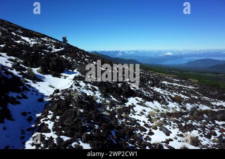 La silhouette del viaggiatore si erge in lontananza sul pendio buio del vulcano Villarrica ricoperto di neve fresca, cielo azzurro, Trekking Villarrica traverse, Vil Foto Stock