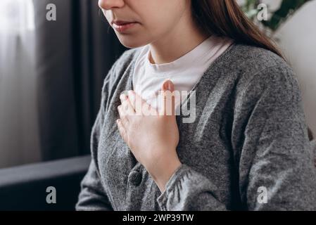 Primo piano di una giovane donna turbata che sente un forte dolore al petto, tormenta il dolore, seduta da sola sul divano di casa. Minaccia di attacco di cuore. Le malattie sono frequenti Foto Stock
