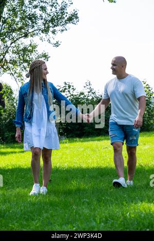 Una coppia sorridente cammina mano nella mano in un parco soleggiato, trasudando calore e connessione. L'immagine evoca temi di amore, compagnia e relax all'aperto Foto Stock