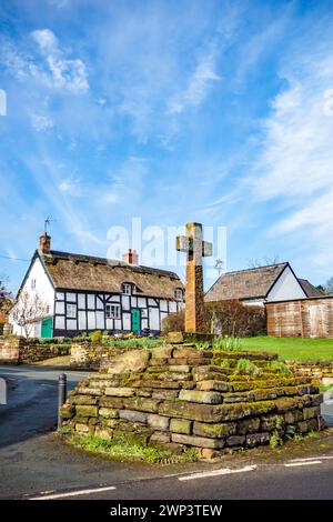 Croce in pietra nel pittoresco e idilliaco villaggio Cheshire di Eaton di Tarporley con un cottage bianco e nero con tetto in paglia in legno Foto Stock