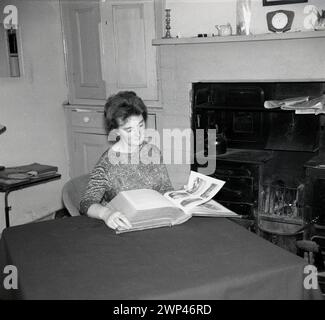 Anni '1960, storica, una signora seduta su un tavolo coperto di stoffa in una cucina di campagna che guarda attraverso un grande e antico libro illustrato, Inghilterra, Regno Unito. All'interno del grande camino, una gamma in ghisa o una stufa e un fuoco aperto. Foto Stock