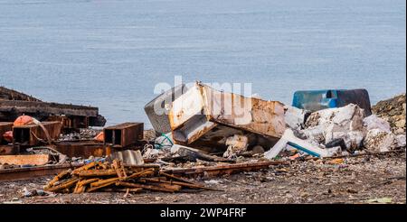 Vecchio frigorifero in metallo arrugginito insieme ad altri rifiuti e detriti che giacevano a terra con l'oceano sullo sfondo in un cantiere navale abbandonato Foto Stock