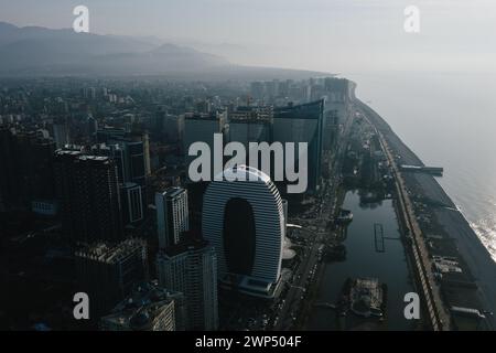 Vista aerea del moderno skyline di Batumi sullo sfondo di una montagna, illuminata dal sole del mattino, che si riflette sugli edifici e sull'oceano. Foto Stock