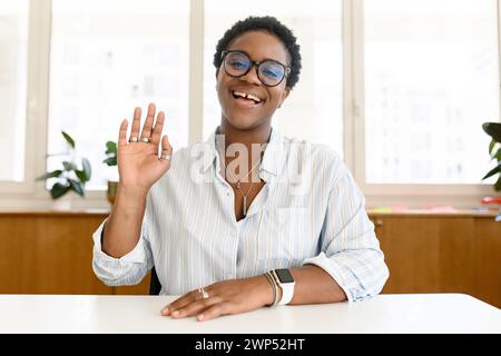 La giovane candidata afroamericana ha coinvolto un colloquio online o una videochiamata, un'insegnante online seduto alla scrivania e guardato e salutato la telecamera, salutando. Caccia al lavoro, concetto di e-learning Foto Stock