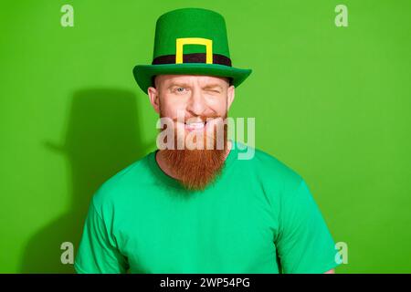 Foto di un uomo seducente e cool con t-shirt con barba allo zenzero e un occhio lampeggiante nella giornata partric isolato su sfondo di colore verde Foto Stock