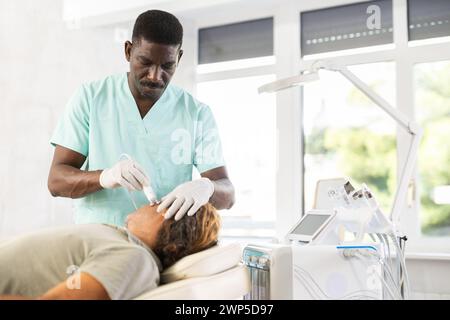 Cosmetologo afroamericano che esegue la procedura di radiofrequenza facciale sull'uomo Foto Stock