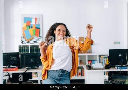 Emozioni gioiose. Allegra ragazza ispanica o brasiliana, dipendente di un ufficio, ballando con gioia in un moderno spazio ufficio, ha ottenuto una promozione, ha chiuso un buon affare, si rallegra alla fine della giornata lavorativa Foto Stock