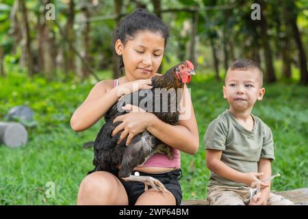 una bruna latina che tiene in mano una gallina nera mentre la accarezza con il fratellino seduto accanto a lei. Foto Stock