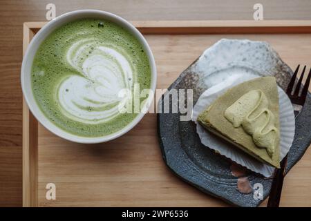 Un latte matcha cremoso e frizzante con latte art, servito in una ciotola di ceramica insieme a una fetta di torta matcha multistrato, presentata su un piatto di ceramica. Foto Stock