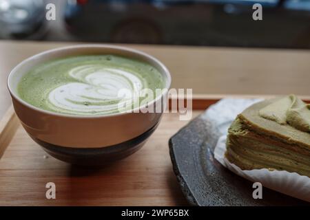 Un latte matcha cremoso e frizzante con latte art, servito in una ciotola di ceramica insieme a una fetta di torta matcha multistrato, presentata su un piatto di ceramica. Foto Stock