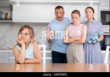 Una triste ragazza prescolare siede a tavola mentre i genitori e la sorella maggiore si rimproverano Foto Stock