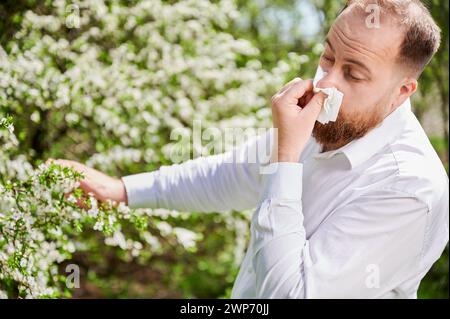 Uomo allergico che soffre di allergia stagionale in primavera nel giardino fiorito in primavera. Giovane che starnutisce e soffia il naso con fazzoletto nasale davanti all'albero in fiore. Concetto di allergia primaverile Foto Stock