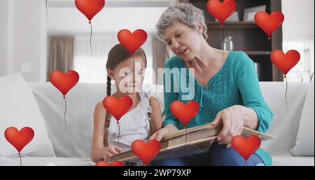 Immagine di palloncini cardiaci sopra la nonna caucasica felice e la nipote che leggono a casa Foto Stock