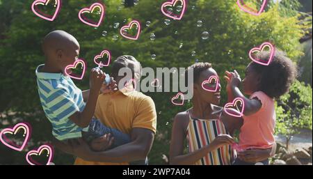 Immagine di cuori su una felice famiglia afroamericana che produce bolle di sapone in giardino Foto Stock