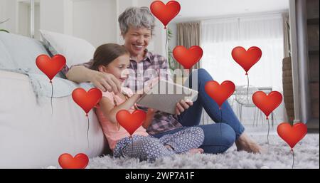 Immagine di palloncini cardiaci sulla nonna caucasica felice e la nipote che usano la tavoletta a casa Foto Stock