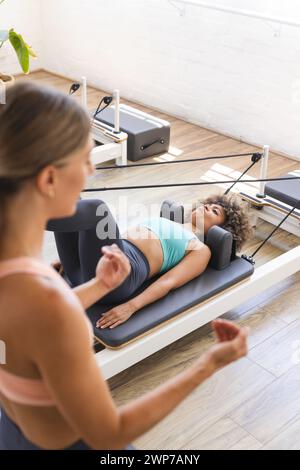 La giovane donna caucasica istruisce una donna birazziale sul riformatore di Pilates Foto Stock