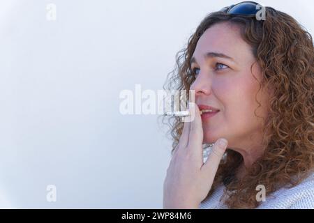 attraente donna di profilo dagli occhi blu e dai capelli ricci che fuma una sigaretta con sfondo bianco e spazio per le copie Foto Stock