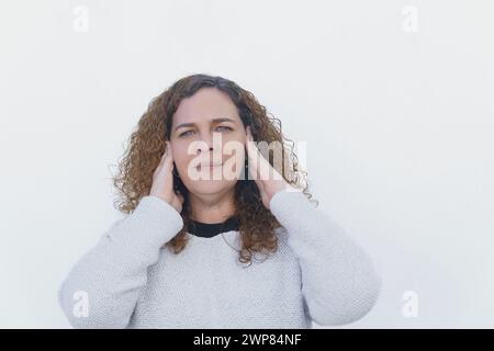 giovane donna attraente con lunghi capelli ricci e occhi blu copre le orecchie con le mani, con sfondo bianco e spazio di copia Foto Stock