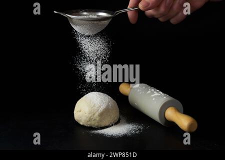 una donna con un setaccio setacciato fa la farina su un impasto di pane accanto a un mattarello isolato su uno sfondo nero e copia spazio Foto Stock