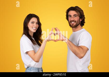 Giovane coppia radiosa che crea una forma a cuore con le mani, sorridendo ampiamente in t-shirt bianche Foto Stock