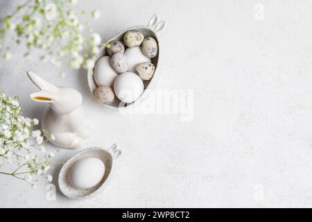 Easter card o invito bianco monocromatico. Due ciotole in marmo figurato con orecchie piene di uova di colore naturale, coniglietto in ceramica, fiori di Gypsophila Foto Stock