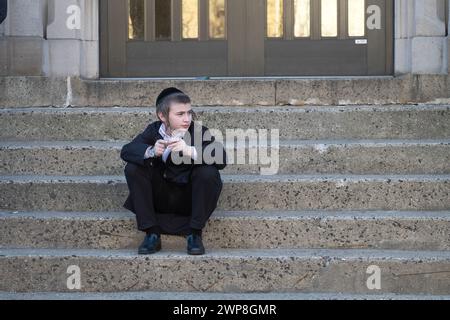 In pausa, un giovane ebreo ortodosso siede sui passi della scuola persi nei suoi pensieri. A Brooklyn, New York, marzo 2024. Foto Stock
