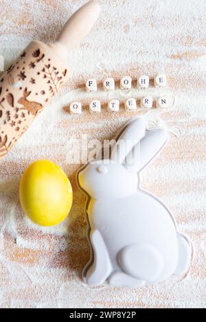 3 marzo 2024: Buona Pasqua, testo su cubetti di legno accanto a una teglia da forno per coniglietto pasquale su una superficie di legno cosparsa di farina *** Frohe Ostern, Text auf Holzwürfeln neben einer Osterhasen Backform auf einer mit Mehl bestreuten Holzfläche Foto Stock