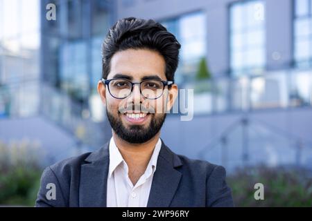 Un giovane uomo d'affari indiano con un abito affilato si erge tranquillamente all'aperto con un paesaggio urbano sullo sfondo. Ideale per temi di diversità aziendale. Foto Stock