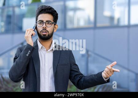 Giovane uomo d'affari professionista in costume che parla con il suo smartphone fuori da un moderno edificio per uffici. Foto Stock