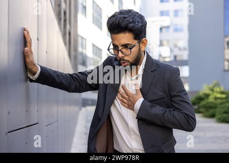 Un uomo professionista in una tuta da lavoro si ferma per un momento fuori, riposando contro un muro con una mano sul cuore, suggerendo contemplazione, sincerità o un concetto di salute. Foto Stock