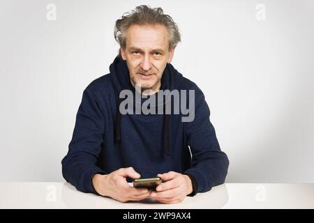 Uomo anziano con cellulare seduto su una camicia con cappuccio al tavolo Foto Stock