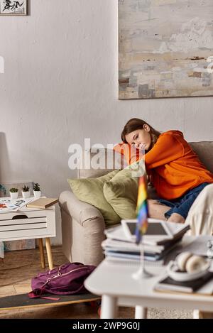 la malinconica ragazza adolescente con cappuccio si siede sul divano con un aspetto lontano, con una bandiera lgbtq sfocata in primo piano Foto Stock