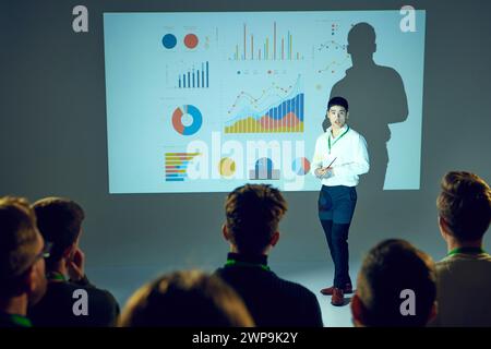 Uomo asiatico, uomo d'affari che tiene conferenze al seminario di affari con partecipanti focalizzati sulle infografiche visualizzate sullo schermo dietro di lui. Foto Stock