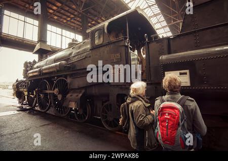 Due donne che guardano la vecchia locomotiva a vapore kkStB 310,23 nel deposito dei treni. L'aria all'interno del capannone del treno è piena di fumo nebbioso. Immagine con tonalità marrone calde. Foto Stock