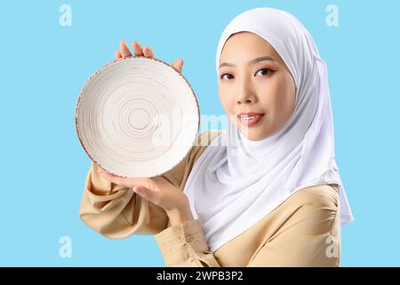 Giovane donna asiatica in hijab con piatto su sfondo blu Foto Stock